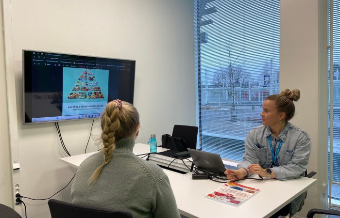 Kaksi ihmistä istuu pöydän ääressä. Seinällä televisiossa ruokapyramidi.