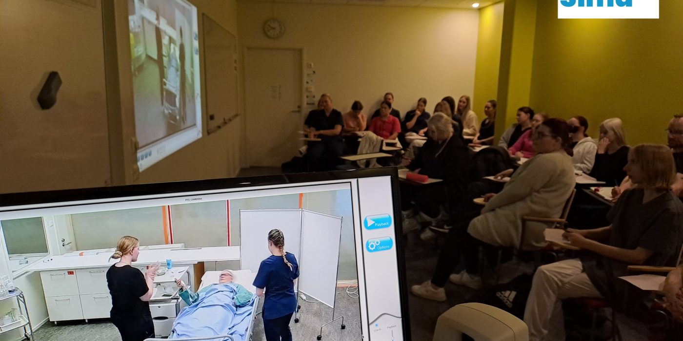 Pimennetyssä luokassa ihmiset katsovat projektorikuvaa simulaatiotilanteesta, jossa hoitotyön opiskelijat hoitavat hoitotyön simulaationukkea.