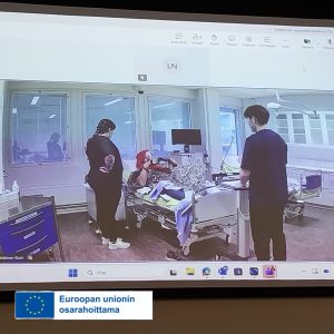 Kuva simulaatiotilanteesta simulaatiomonitorin kautta. Simulaatiotilanteena potilastilanne jossa potilas sängyllä ja kaksi hoitajaa toimii vieressä.