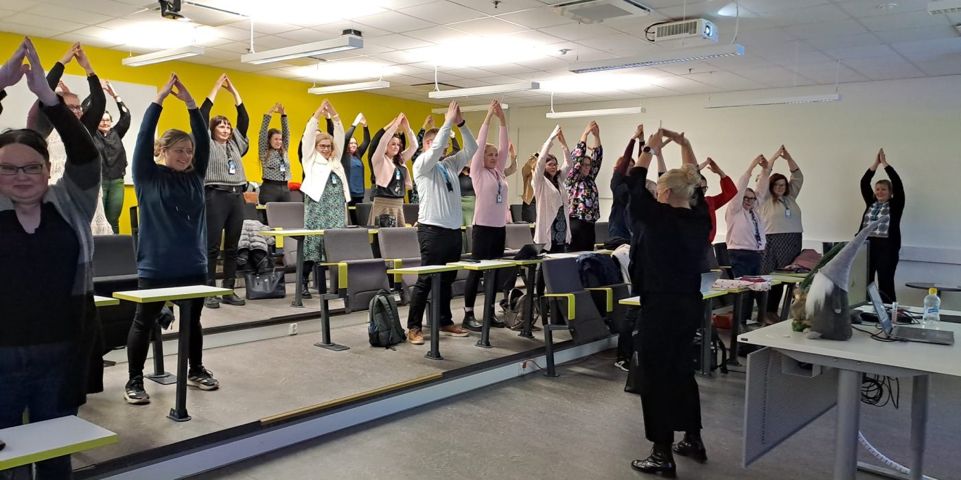 SAMKin Porin kampuksen auditoriossa yleisö seisoo kädet ylhäällä. Edessä ohjaaja niin ikään kädet ylhäällä ohjaamassa jumppahetkeä.