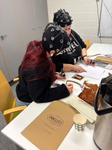 Kaksi henkilöä työskentelevät pöydän ääressä pakopeliä erilaisilla papereilla. Päässään heillä on eeg-myssyt.