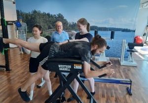 Opiskelijat Julianna Pakkanen (vasemmalla) ja Riina Urpilainen ohjaamassa asiakas Joonas Saarelaa selkälaitteessa fysioterapiakeskus Fyysingissä toteutetussa MUNDO-harjoittelussa. Taustalla tilannetta seuraa Nina Nieminen, yrityksen fysioterapiavastaava ja harjoitteluohjaaja.